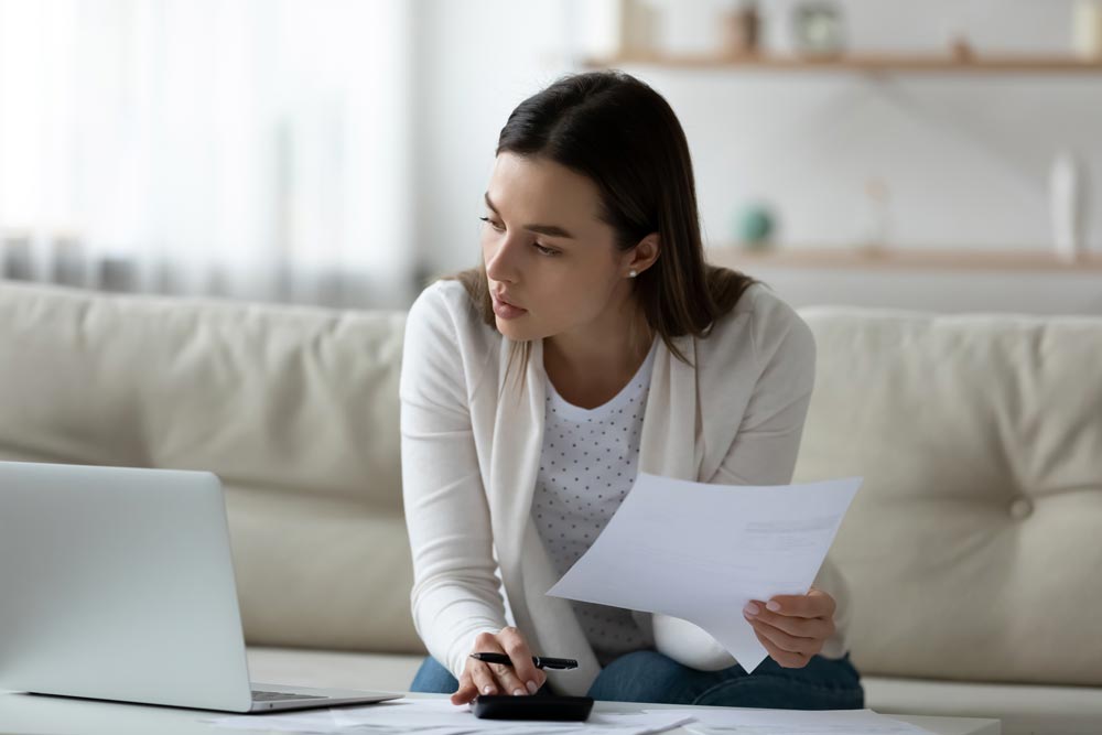 Singh Associates Individual Tax Returns Individual taxes - Woman sitting on couch working on laptop and holding paperwork.